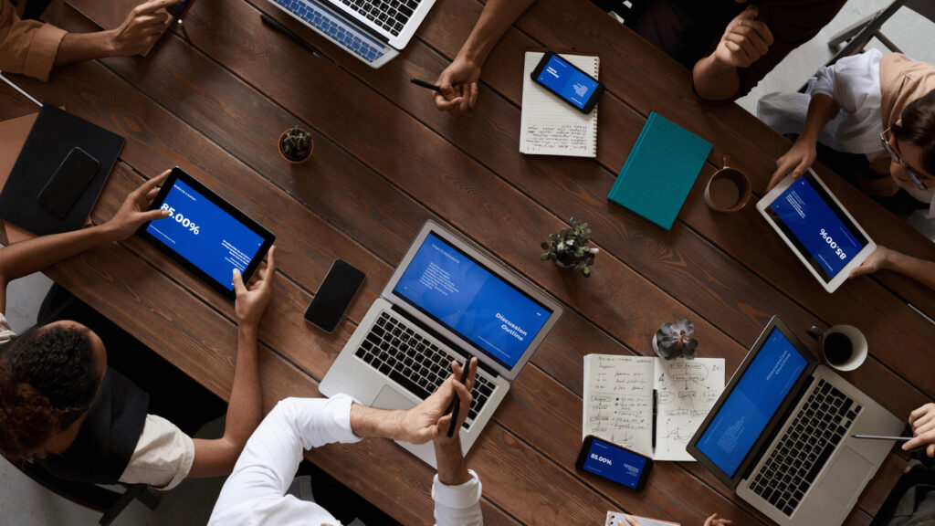 A group of people gathered around a wooden table with laptops, tablets, and phones, engaged in a meeting for How to Write a Meeting Agenda.