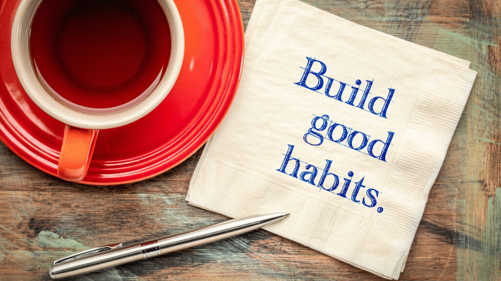 A white napkin with the words "Build good habits, “ written in blue text, lies next to a red teacup and saucer and a silver pen on a wooden table, symbolizing a mindful work environment and productivity.