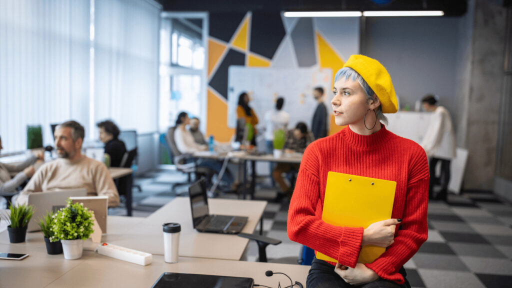 A person wearing a red sweater and a yellow beret holds a yellow clipboard in a modern office setting with other people working in the background, deciding between a hot desk vs. a dedicated desk.
